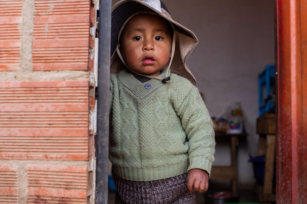A little boy in a green sweater and floppy hat looks at the camera.